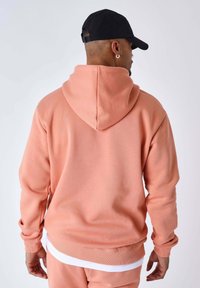 Sudadera con capucha rosa con cordón, puños y dobladillo acanalados, llevada sobre una capa blanca. Textura lisa, ajuste casual, vista desde atrás.