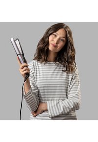 Piastra per capelli con involucro grigio metallico, designSlim e bordi arrotondati. Il modello la tiene con il cavo attaccato, indossando una maglietta a righe grigie chiare.