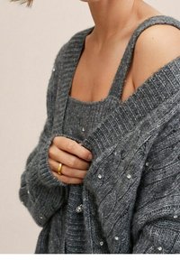 Grauer gestrickter Cardigan mit gerippter Textur, verziert mit kleinen Kristallen. Er hat einen tiefen V-Ausschnitt, weite Ärmel und ein geschichtetes Design über einem Tanktop.
