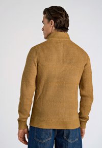 Stickad tröja i gyllengul färg med hög krage och ribbad struktur, med långa ärmar och rak nederkant, kombinerad med blå jeans.