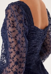 Mörkblå klänning med genomskinliga långärmade ärmar som har intrikata blommönster, rynkad byst och fyrkantig halsringning.