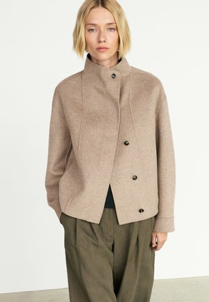Kvinde med kort blondt hår bærer en beige knappet jakke og olivengrønne bukser, stående mod en ensfarvet lys baggrund.