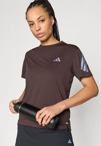 Brązowa sportowa koszulka wykonana z miękkiego materiału, z krótkimi rękawami, z jasnoniebieskim logo i paskami na ramionach. Czarna butelka na wodę trzymana w dłoniach.