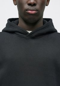 Primo piano della parte inferiore del viso e del collo di una persona che indossa una felpa con cappuccio nera sopra una maglietta bianca, su uno sfondo chiaro e uniforme.