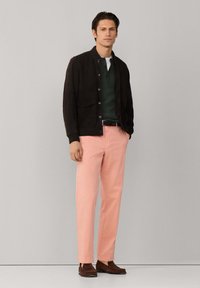 Homme debout contre un mur uni portant une veste noire, une chemise vert foncé, un pantalon rose et des mocassins marron, les mains dans les poches.