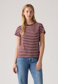 T-shirt rayé bordeaux et blanc avec un col rond et des manches courtes, arborant un petit logo sur la poitrine gauche. Porté avec un jean bleu.