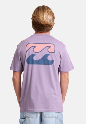 Persona con capelli biondi che indossa una maglietta lavanda con un motivo a onda stilizzata rosa e blu sulla schiena e pantaloncini di jeans celesti.