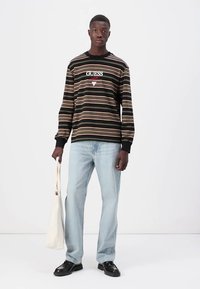 Svart langermet stripet skjorte med rød og hvit logo, sammen med lyseblå jeans og svarte sko. Holder en kremfarget toteveske.