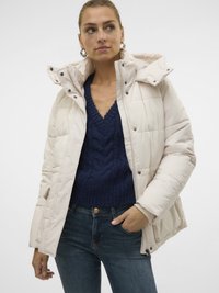 Ženska, ki nosi kremasto odejano jakno čez mornarsko modro pulover z V-izrezom in modre kavbojke, z eno roko v žepu, ki gleda vstran.