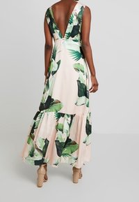 Robe longue en satin, avec un dos en V, un motif de feuilles tropicales vertes sur un fond rose clair, une jupe à volants, et une longueur jusqu'aux chevilles.