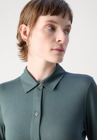 Polo vert avec col, fermeture à boutons et texture lisse. Présente un design ajusté et un éclat subtil, sans motifs visibles.