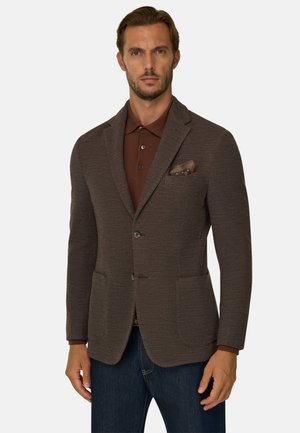 Homme portant un blazer marron texturé avec deux poches avant, une chemise marron à col et un jean bleu foncé sur un fond uni.