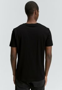 Musta lyhyttaskuinen t-paita, pyöreä pääntie, sileä rakenne, vartalonmyötäinen muotoilu, esitetty takaa. Yhdistetty tummiin farkkushortseihin.