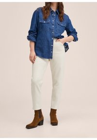 Camisa de botones de mezclilla en azul oscuro, con dos bolsillos en el pecho. Combinada con pantalones beige claros y botas de tobillo marrones con tacón cuadrado.