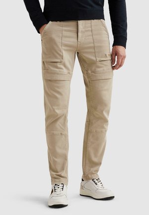Hombre con pantalones cargo beige con bolsillos delanteros de parche y zapatillas blancas, de pie con las manos cerca de los bolsillos.