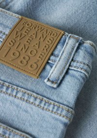 Gaiši zils džinsa audums ar teksturētu virsmu. Ir brūna reljefa ādas uzlika ar uzrakstu "FORWARD JEANS WEAR SINCE 1986".