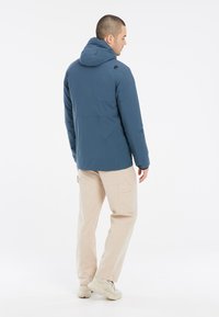 Giacca isolante blu con cappuccio, caratterizzata da un design elegante e una superficie liscia. Abbinata a pantaloni cargo beige e scarpe di colore chiaro.