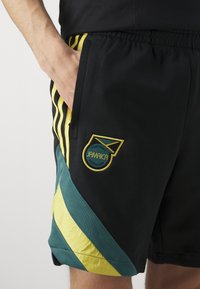 Pantalones cortos negros con rayas amarillas y acentos en azul verdoso, que cuentan con el logo de la Federación de Fútbol de Jamaica. La tela parece suave y tiene una cinturilla elástica.