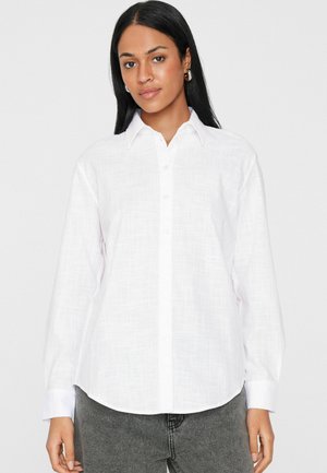 Frau mit glatten schwarzen Haaren, die silberne Creolen, ein strukturiertes weißes Hemd mit Knöpfen und graue Jeans trägt, stehend vor einem weißen Hintergrund.