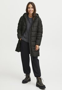 Giacca puffa verde scuro con cappuccio, design trapuntato, zip frontale e tasche laterali, indossata sopra un maglione grigio e pantaloni neri.