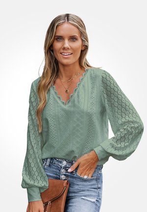 Vrouw die een lichtgroene kanten blouse met opgeblazen mouwen draagt, gecombineerd met high-waisted jeans met knoopsluiting en een bruine clutch vasthoudt.