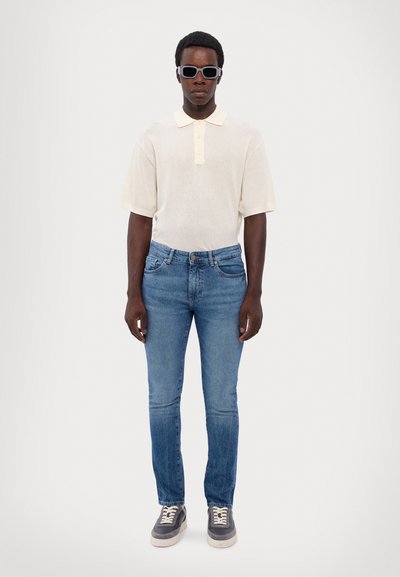 Tejfehér hálós póló rövid ujjakkal, világoskék, slim-fit farmernadrág, és szürke sportcipő fehér fűzővel és fehér talppal.