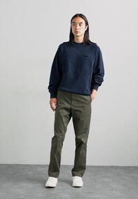 Sudadera navy con mangas raglán, pantalones verde oliva y zapatillas blancas. La sudadera presenta un pequeño detalle de bolsillo en la parte frontal.