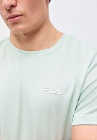 Κοντινό πλάνο ενός ατόμου που φοράει ένα ανοιχτό μέντα πράσινο μπλουζάκι FILA με στρογγυλή λαιμόκοψη, εμφανίζοντας μέρος του προσώπου και του άνω κορμού.