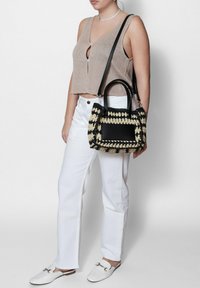 Borsa a tracolla intrecciata nera e crema con un accento rettangolare in pelle nera, manici corti e una lunga tracolla, abbinata a un top beige corto e pantaloni bianchi.