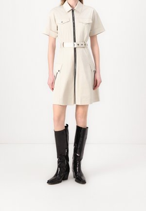 Woman wearing a beige belted shirt dress with front zipper, short sleeves, and black knee-high heeled boots against a plain white background.