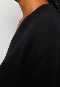 Maglione nero con scollatura a V profonda, tessuto a maglia testurizzata e dettagli a coste lungo le spalle. La vista laterale mostra la vestibilità e la caduta.