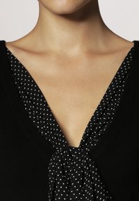 Czarny sweter z dekoltem w kształcie litery V, z białym, marszczonym materiałem w kropki przy dekolcie, tworzącym elegancki szczegół w formie węzła.