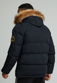 Schwarze Puffy-Jacke mit einer mit Fell gefütterten Kapuze, gesteppter Optik und einem bestickten Patch auf der linken Schulter. Verfügt über einen robusten, strukturierten Stoff.