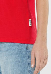 Red cotton T-shirt featuring a round neck and short sleeves. A woven label with "SENSES" is attached to the seam. Light blue denim visible.
