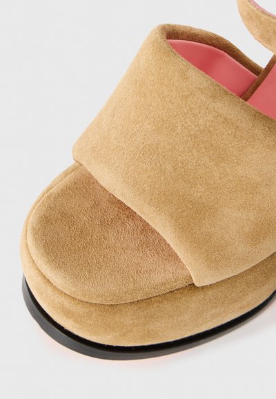 Beige mockaskalsong med en bred rem över framsidan, mjuk textur, rosa inredning och en svart sula. Rundad tådesign.