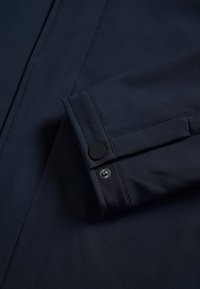 Manche de manteau bleu marine foncé avec des lignes de couture visibles et une fermeture pression noire au niveau du poignet.