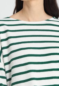 Nainen, jolla on yllään tekstuurilla varustettu valkoinen neulepaita, jossa on tasaisesti sijoitettuja vaaleanvihreitä vaakaraitoja ja leveä pyöreä kaula-aukko.