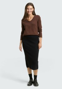 Brauner V-Ausschnitt-Pullover kombiniert mit einem schwarzen, knielangen Rock. Das Modell trägt schwarze Socken und Schnürschuhe und steht vor einem neutralen Hintergrund.