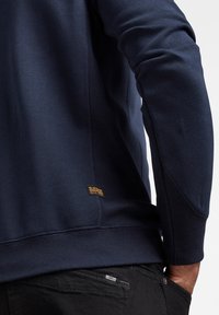 Gros plan d'une personne portant une chemise bleu marine à manches longues avec un petit patch G-Star et un pantalon noir, main reposant dans la poche arrière.
