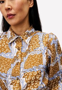 Braun gemustertes Hemd mit blauen Blumen- und Leopardenmotiven, Knopfleiste, und einem normalen Kragen mit leicht puffenden Ärmeln.