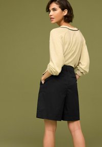 Pull beige avec des manches froncées et short noir. Le short a des poches latérales et une coupe structurée. L'arrière-plan est d'un vert olive uni.