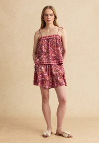 Mujer de pie con las manos en los bolsillos, lleva un top sin mangas rosa con estampado de cachemira y pantalones cortos a juego, con sandalias de ganchillo color crema.