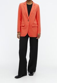 Blazer naranja con diseño estructurado, cierre de un solo botón, dos bolsillos frontales, usado sobre una blusa negra y combinado con pantalones de pierna ancha negros.
