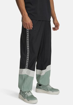 Uomo che indossa pantaloni a blocchi di colore nero, bianco e verde con una striscia a catena bianca e scarpe da ginnastica verdi, in piedi contro uno sfondo uniforme.