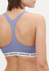 Lavendelfärgad sportbehå med racerback-design, prydd med ett vitt band med "BJÖRN BORG" i svart text. Mjuk och slät tygkänsla.