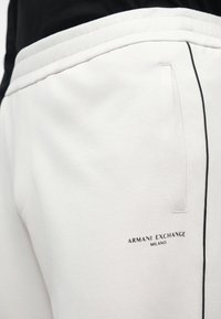 Biele jogger nohavice vyrobené z mäkkého materiálu, s čiernym logom "ARMANI EXCHANGE MILANO" a kontrastným čiernym lemovaním po stranách.
