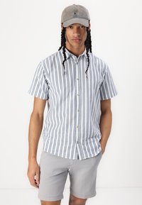 Chemise à manches courtes avec des rayures verticales bleu et blanc, associée à un short gris clair. Le modèle porte une casquette gris clair.
