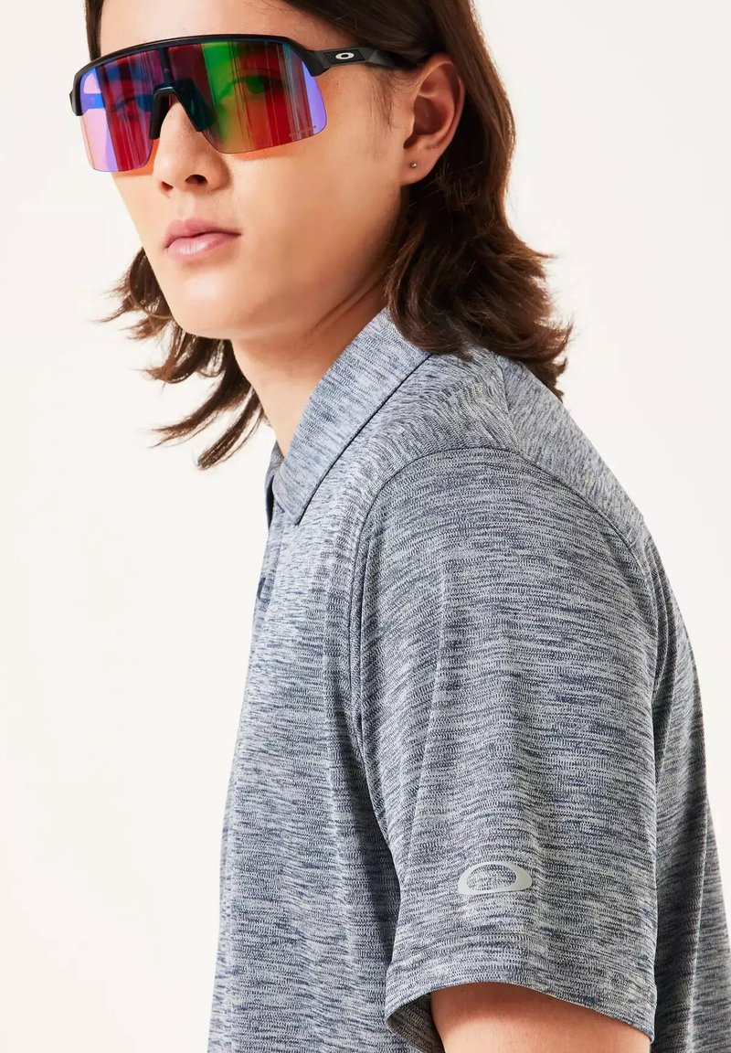 Persona con cabello hasta los hombros, vistiendo una camiseta polo gris con textura y grandes gafas de sol deportivas reflectantes, mirando ligeramente hacia la izquierda.