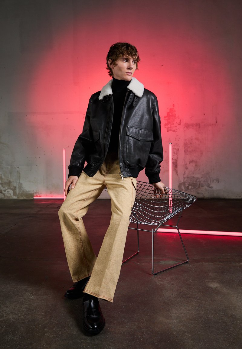 Black leather jacket with white shearling collar, paired with beige wide-leg pants. Model seated on a wire chair against a pink backdrop.