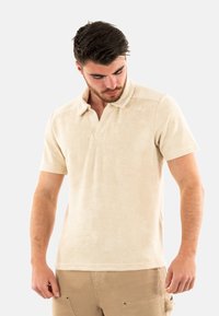 Polo shirt beige a maniche corte in morbido tessuto strutturato. Presenta un colletto classico e un logo discreto sul petto. Vestibilità casual.